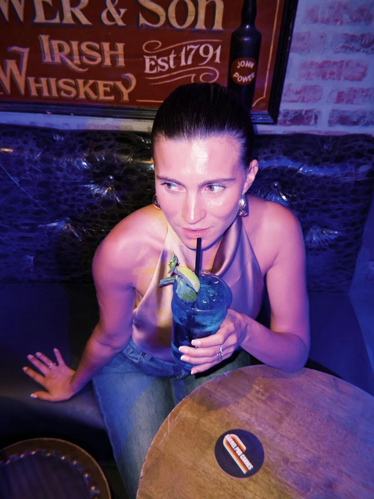 Une cliente penchée sur un tonneau, cocktail bleu à deux pailles, sous l'enseigne Power & Son Irish Whiskey